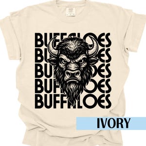 Może przedstawiać: Kremowa koszulka z czarną grafiką głowy bawołu i słowem "BUFFALOES" ułożonym pionowo po lewej stronie. Słowo "IVORY" jest wyświetlane w jasnoniebieskim prostokącie na dole.