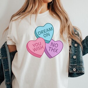 Puede incluir: Camiseta blanca con tres diseños de caramelos en forma de corazón. Los corazones dicen "Dream On", "You Wish" y "No Thx".