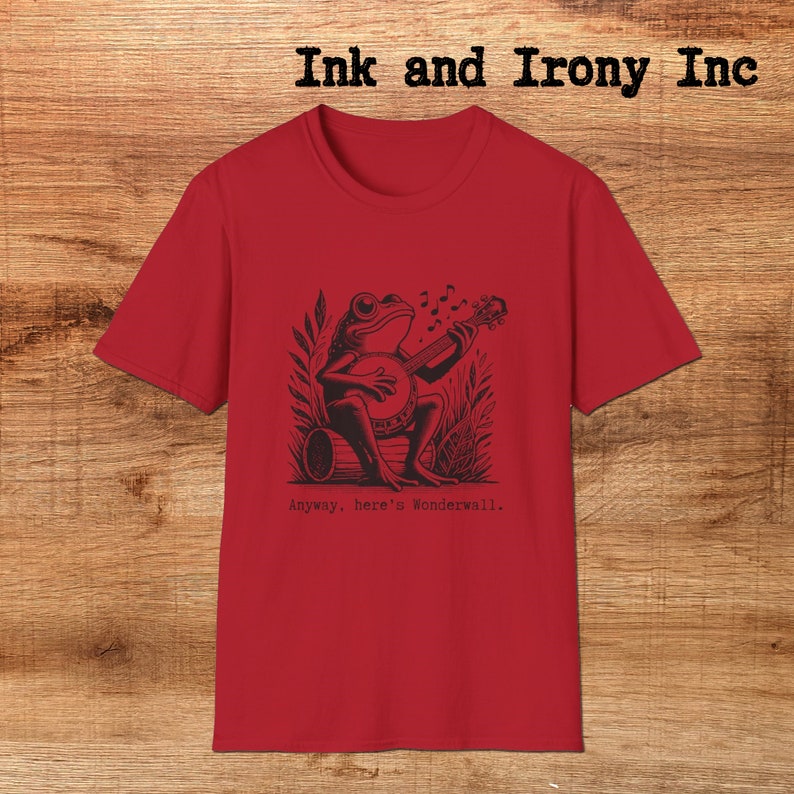 May include: Red t-shirt with a black and white illustration of a frog playing a banjo. The text "Anyway, here's Wonderwall." is printed below the illustration.
