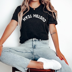 Puede incluir: Camiseta gris oscuro con el texto "CUSTOM TEAM" en blanco, arqueado sobre el pecho. La camiseta se combina con vaqueros azul claro y zapatos blancos sin cordones. La persona está sentada en un taburete de madera.