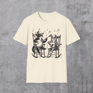 May include: A cream-colored t-shirt with a black and white illustration of two cats wearing cowboy hats and playing musical instruments. One cat plays a banjo and the other plays a guitar. The cats are sitting in rocking chairs.