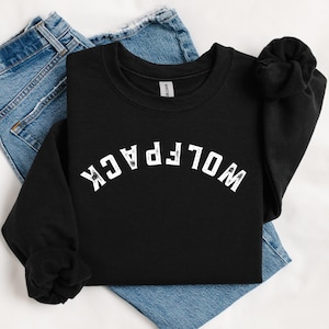 Puede incluir: Una sudadera negra con la palabra "WOLFPACK" en blanco, con letras arqueadas en la parte delantera. La sudadera está doblada sobre un par de vaqueros azules. La etiqueta de la marca Gildan es visible en el interior del cuello.