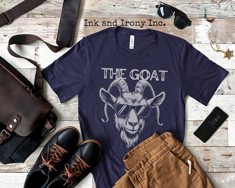 May include: A navy blue t-shirt featuring a graphic of a goat wearing sunglasses and the text "THE GOAT" printed on the front.