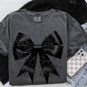May include: Dark gray t-shirt featuring a large black bow design covered in spiderwebs and spiders. The bow is centered on the shirt. The shirt is folded with a pair of blue jeans and a phone case with a checkered pattern.