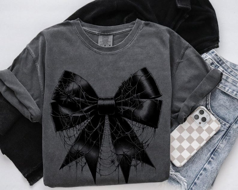 May include: Dark gray t-shirt featuring a large black bow design covered in spiderwebs and spiders. The bow is centered on the shirt. The shirt is folded with a pair of blue jeans and a phone case with a checkered pattern.