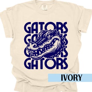 Peut inclure: T-shirt ivoire avec le mot "GATORS" répété trois fois et un dessin d'alligator bleu marine. Le t-shirt a un col rond et des manches courtes. Le mot "IVORY" est affiché en bas.
