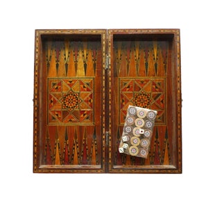 Puede incluir: Un tablero de backgammon de madera con intrincados diseños de incrustaciones en tonos marrones, rojos y dorados. El tablero está abierto y una pila de fichas de juego es visible en el lado derecho del tablero.