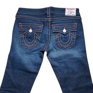 May include: Dark wash denim jeans with signature back pocket stitching and a True Religion label. The jeans feature orange stitching and silver hardware. The back pockets have a horseshoe design.