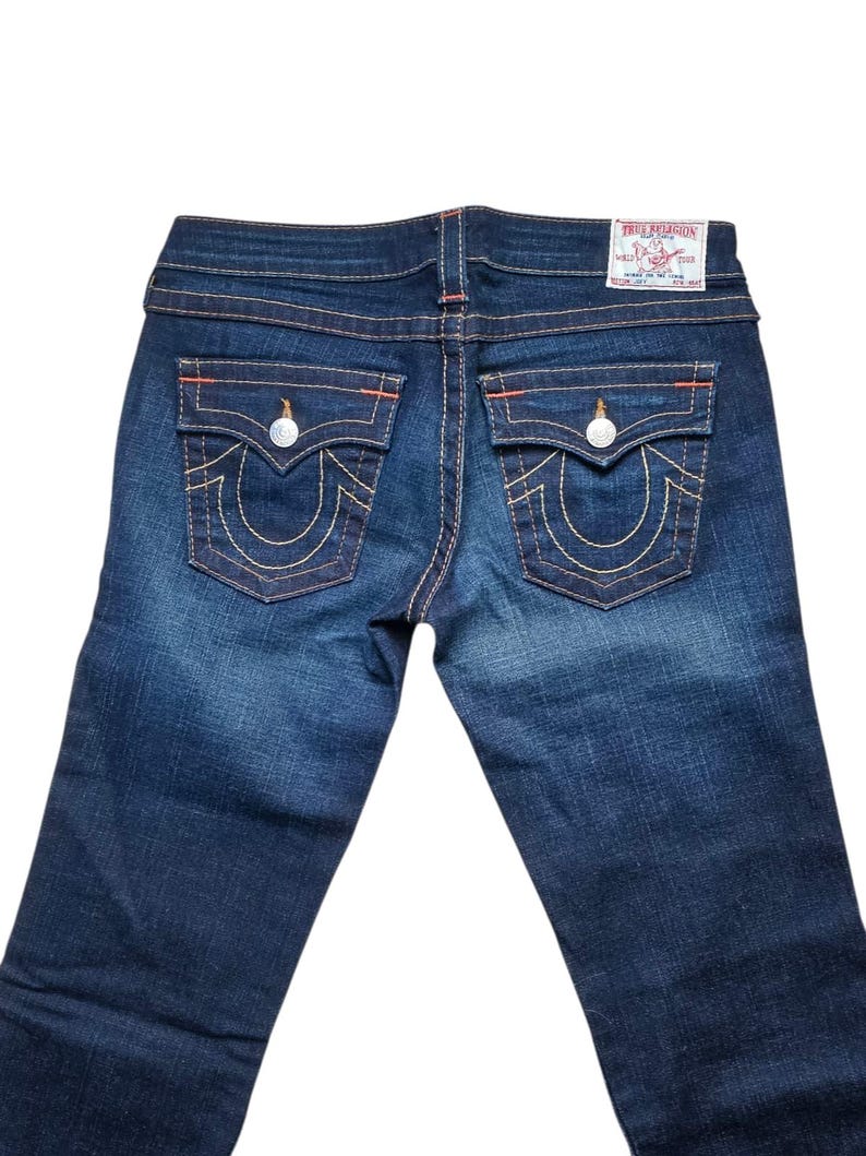 May include: Dark wash denim jeans with signature back pocket stitching and a True Religion label. The jeans feature orange stitching and silver hardware. The back pockets have a horseshoe design.