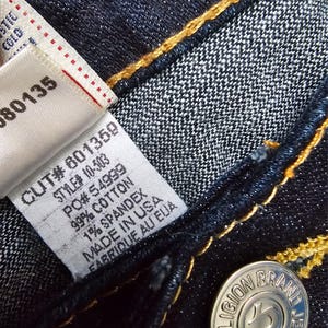 May include: Close-up of dark blue denim jeans with yellow stitching. A white tag displays the text "CUT# 601359, STYLE# 10-503, 99% COTTON, 1% SPANDEX, MADE IN USA, FABRIQUE AU EUA." A silver button reads "TRUE RELIGION BRAND JEANS."