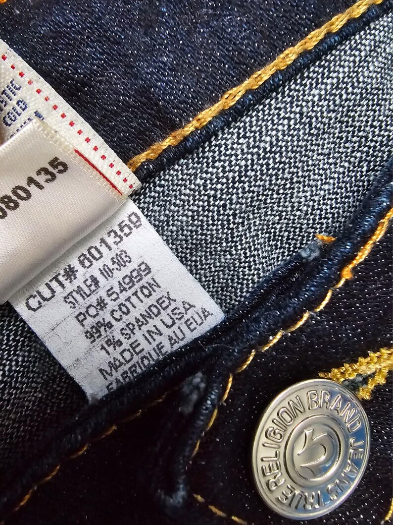 May include: Close-up of dark blue denim jeans with yellow stitching. A white tag displays the text "CUT# 601359, STYLE# 10-503, 99% COTTON, 1% SPANDEX, MADE IN USA, FABRIQUE AU EUA." A silver button reads "TRUE RELIGION BRAND JEANS."