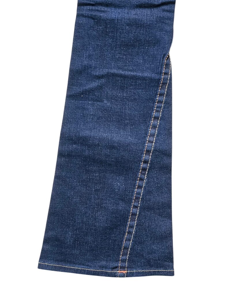 May include: Close-up of a pair of dark blue denim jeans. The jeans have a flared leg and a visible seam with white stitching running down the side. The denim has a classic, textured appearance.