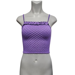 May include: A purple crop top with thin straps and a ruffled neckline. The top has a textured, woven pattern and is displayed on a mannequin. The garment is a solid color and appears to be made of a stretchy material.