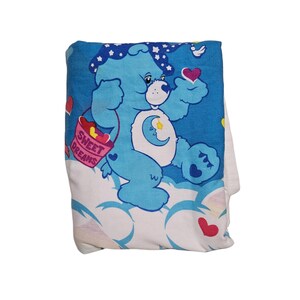 May include: A blue Care Bear with a star on its head and a crescent moon on its belly is holding a pink heart. The bear is standing on a white cloud with the words "Sweet Dreams" on a pink banner. The background is blue with white stars and pink hearts.