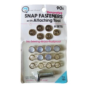 Puede incluir: Un paquete de seis juegos de broches de presión decorativos de latón inoxidable con una herramienta de fijación. El empaque blanco presenta el texto Decorative SNAP FASTENERS with Attaching Tool, 6 SETS, No Sewing e ITEM 191. Los broches son dorados.