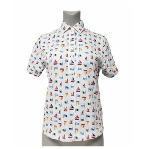 May include: White short-sleeve shirt with a sailboat pattern. The shirt features a classic collar, a button-down placket, and rolled-up sleeves. The sailboats are in red, blue, yellow, and green. The shirt is displayed on a mannequin.
