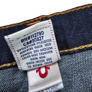 May include: Close-up of a dark blue denim fabric with a white tag. The tag has text including "RN#112790" and "CA#30427", along with washing instructions. A smaller tag features a red horseshoe logo and "MADE IN USA".