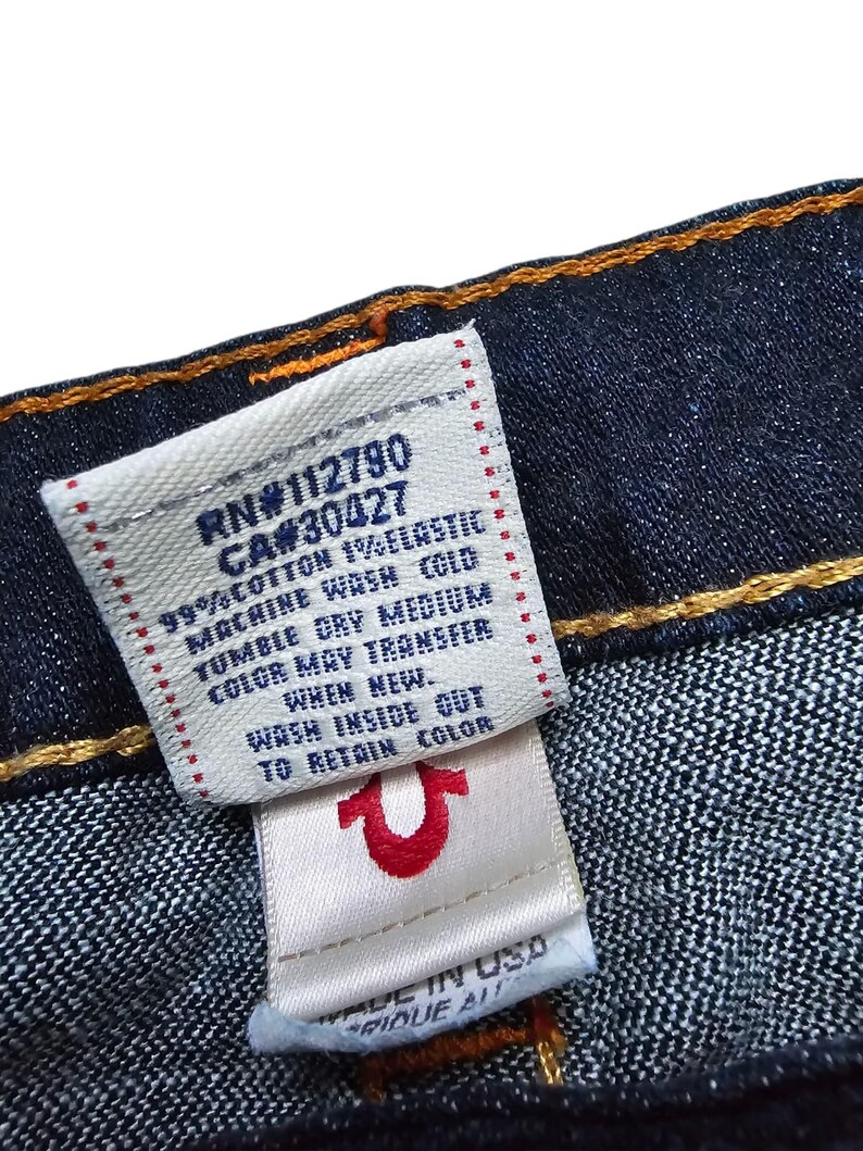May include: Close-up of a dark blue denim fabric with a white tag. The tag has text including "RN#112790" and "CA#30427", along with washing instructions. A smaller tag features a red horseshoe logo and "MADE IN USA".
