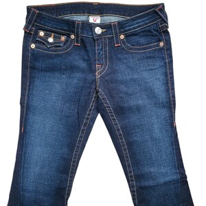 May include: Dark wash denim jeans with a classic five-pocket design. Features include a button and zipper closure, decorative stitching, and a small logo detail. The jeans have a slight flare at the bottom.
