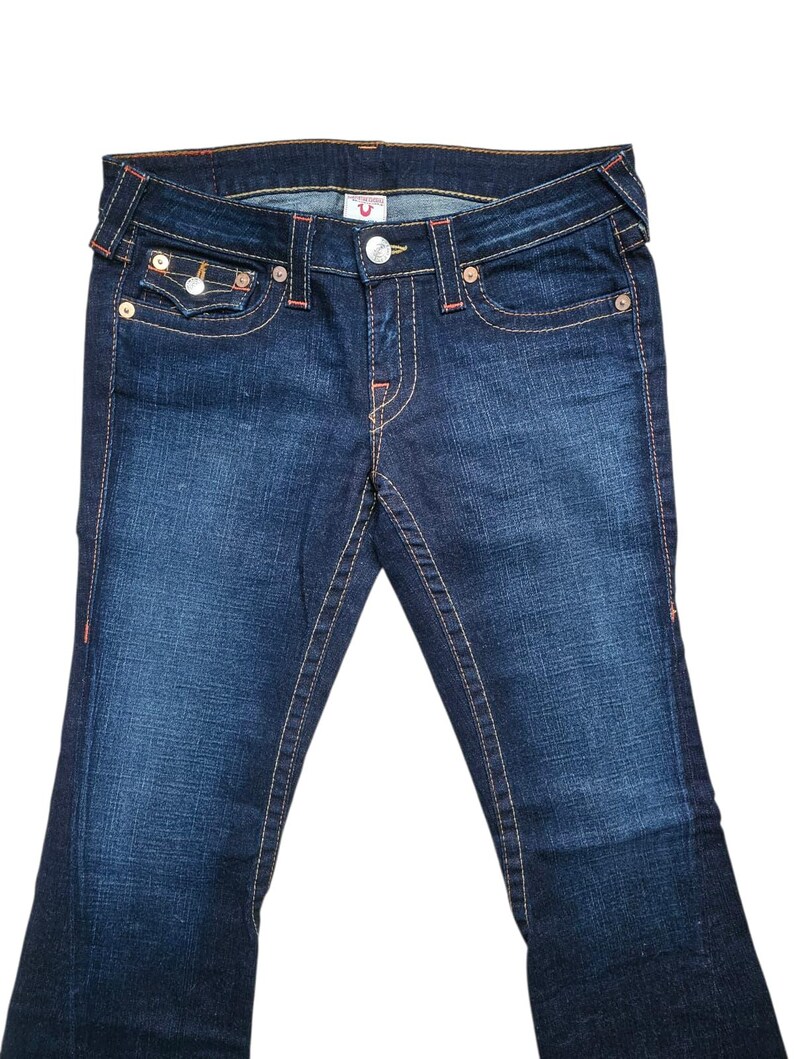 May include: Dark wash denim jeans with a classic five-pocket design. Features include a button and zipper closure, decorative stitching, and a small logo detail. The jeans have a slight flare at the bottom.