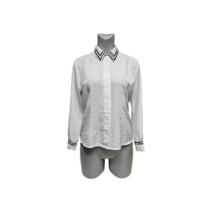 May include: A white long-sleeve shirt with a classic collar and cuffs. The collar and cuffs feature a black and white striped design. The shirt is buttoned down the front and is displayed on a mannequin.