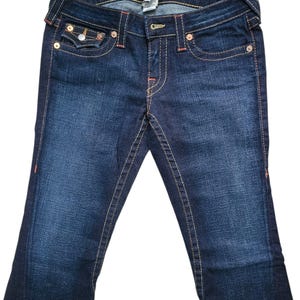 May include: Dark wash denim jeans with a bootcut leg. The jeans feature orange stitching, a button and zipper closure, and classic five-pocket styling. The back pockets have a decorative design. The jeans are a medium rise.
