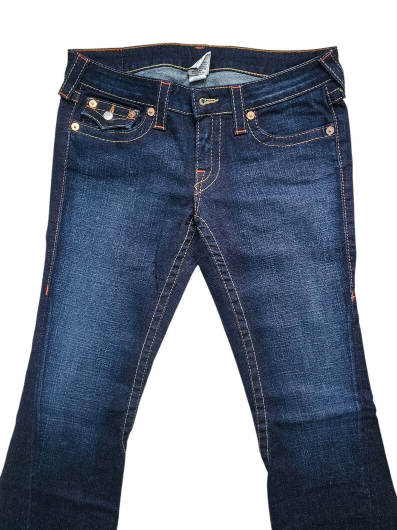 May include: Dark wash denim jeans with a bootcut leg. The jeans feature orange stitching, a button and zipper closure, and classic five-pocket styling. The back pockets have a decorative design. The jeans are a medium rise.
