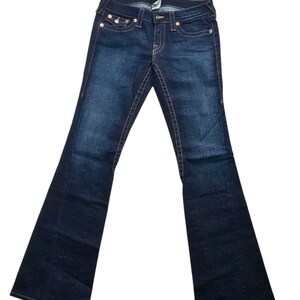 May include: Dark wash denim jeans with a flared leg. The jeans feature classic five-pocket styling, copper-colored stitching, and a button and zipper closure. The back pockets have a decorative stitch design.