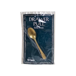 May include: A solid brass drawer pull in the shape of a spoon, packaged in clear plastic. The product is on a dark blue background with the words "DRAWER PULL" and "Solid Brass". The packaging also includes the Pier 1 Imports logo.