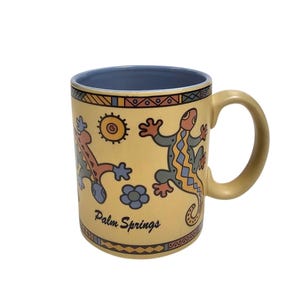 May include: A yellow ceramic mug with a light blue interior and handle. The exterior features a lizard graphic, a sun icon, and flower shapes in orange, blue, and brown. The text Palm Springs is printed in black script on the front of the mug.
