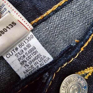 May include: Close-up of dark blue denim jeans with yellow stitching. A white tag displays the text "CUT# 601359, STYLE# 10-503, 99% COTTON, 1% SPANDEX, MADE IN USA, FABRIQUE AU EUA." A silver button reads "TRUE RELIGION BRAND JEANS."