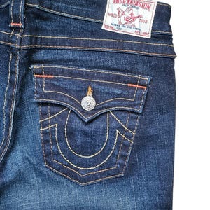 May include: Close-up of dark wash denim jeans. The back pocket features a decorative stitch design and a silver button. The True Religion brand label is visible at the waistband. Orange stitching accents the seams.