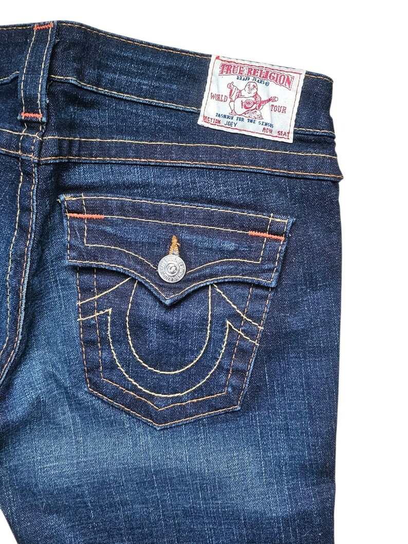 May include: Close-up of dark wash denim jeans. The back pocket features a decorative stitch design and a silver button. The True Religion brand label is visible at the waistband. Orange stitching accents the seams.
