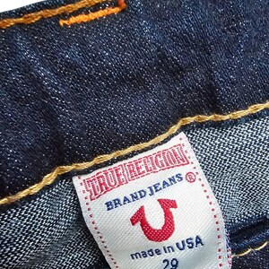 May include: Close-up of dark blue denim jeans with orange stitching. A white label reads "TRUE RELIGION BRAND JEANS" with a red horseshoe logo and "made in USA 29".