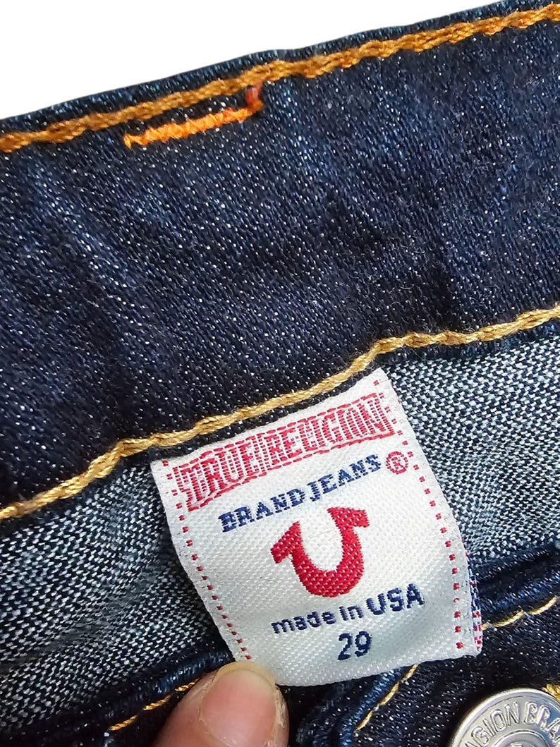 May include: Close-up of dark blue denim jeans with orange stitching. A white label reads "TRUE RELIGION BRAND JEANS" with a red horseshoe logo and "made in USA 29".
