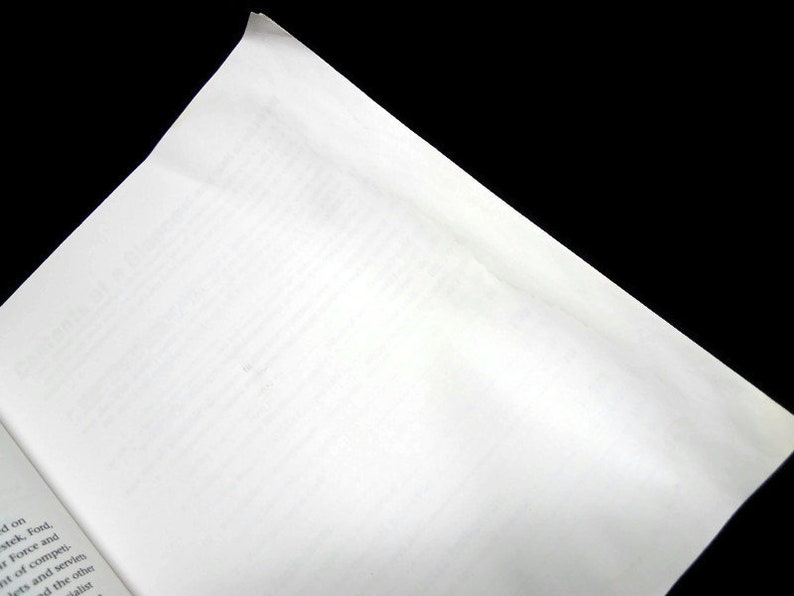 以下が含まれることがあります： 本を開いた状態のクローズアップ。右ページは右上隅に向かって傾いています。ページは白く、左下のページにテキストが見えます。背景は黒です。