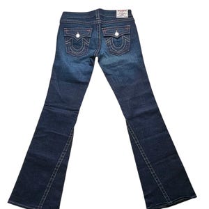 May include: Dark wash denim jeans with a flared leg. The jeans feature white stitching, two back pockets with a signature design, and a small logo patch. The back pockets have a red outline. The jeans are displayed on a white background.