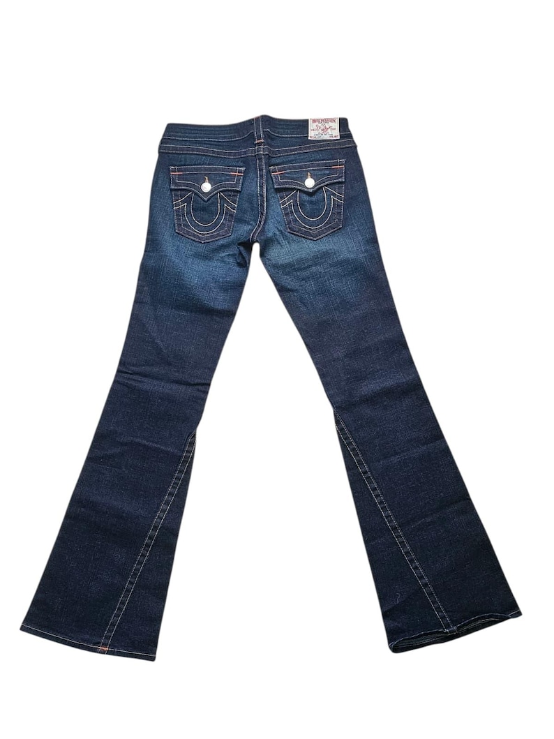 May include: Dark wash denim jeans with a flared leg. The jeans feature white stitching, two back pockets with a signature design, and a small logo patch. The back pockets have a red outline. The jeans are displayed on a white background.