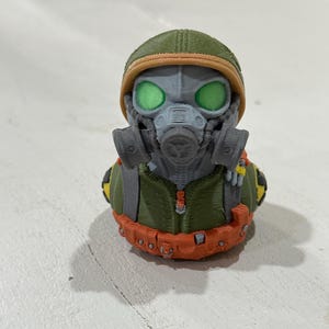 May include: A 3D-printed rubber ducky wearing a gas mask and military helmet. The ducky has green eyes, a gray gas mask, and an olive green uniform. An orange belt wraps around the ducky's midsection.