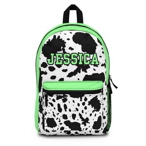 May include: A backpack with a black and white cow print design, with neon green trim. The name "JESSICA" is printed in green block letters on the front. Features a front pocket, side mesh pocket, and black zip.