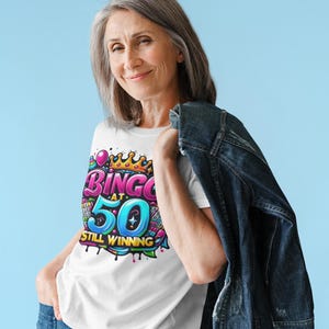 May include: A white t-shirt with a colorful graphic design that says "Bingo at 50 Still Winning". The design features a crown, balloons, and bingo cards.
