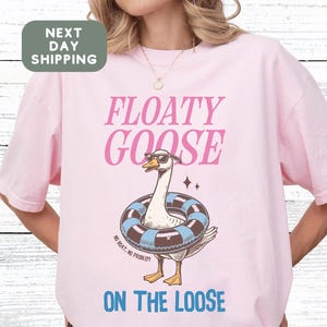 May include: A pale pink t-shirt featuring the text "FLOATY GOOSE" in pink and blue. A cartoon goose wearing sunglasses and a swim ring is in the center. The words "ON THE LOOSE" are below the goose.