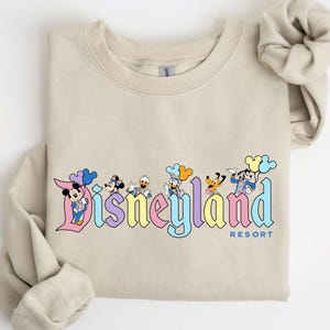 以下が含まれることがあります： ミッキーマウス、ミニーマウス、ドナルドダック、グーフィー、プルート、チップとデールが描かれたカラフルなディズニーランドリゾートのロゴが入ったベージュのトレーナーです。