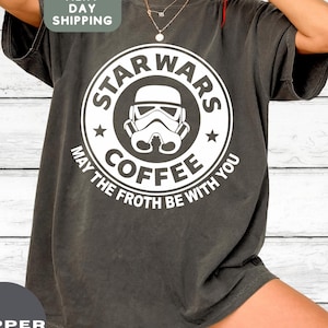 May include: Dark gray t-shirt with a white graphic featuring a Stormtrooper helmet and the words "STAR WARS COFFEE" in a circular design. The text "MAY THE FROTH BE WITH YOU" is also included.