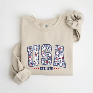 May include: A beige sweatshirt with the word "USA" in a floral design. The letters are outlined in navy blue and filled with a pattern of red and white flowers. Below the word "USA" is the text "EST. 1776" with red stars.