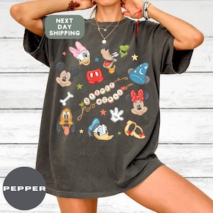 May include: Dark gray t-shirt featuring Disney characters like Mickey Mouse, Minnie Mouse, Donald Duck, and Pluto. The shirt has the words "Disney World" in a colorful design. The shirt also has the text "NEXT DAY SHIPPING".