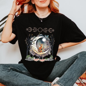 May include: Black t-shirt featuring a whimsical design of a fox sitting on a crescent moon, surrounded by flowers and trees. The design includes phases of the moon across the top. A nature-themed graphic tee.