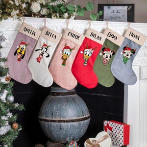 May include: Six Christmas stockings in various colors, each with a different Disney character wearing a Santa hat and scarf. Names are printed above each character: Amelia, Bella, Chloe, Dylan, Emma, and Fiona. The stockings are hanging above a fireplace.