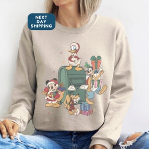May include: Beige sweatshirt featuring a festive graphic of Disney characters Mickey Mouse, Donald Duck, Goofy, and Pluto. The characters are depicted in holiday attire, surrounded by gifts and a mailbox. The sweatshirt has a crew neck and long sleeves.