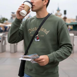 A man wearing a green sweatshirt with the Disney logo is drinking from a coffee cup and holding a map, with a Disney theme park visible in the background.